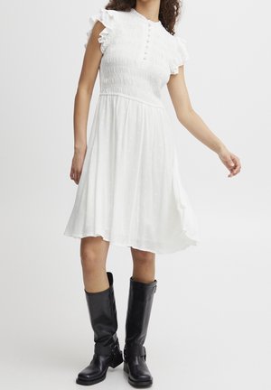 Frau trägt ein weißes knielanges Kleid mit Rüschenärmeln und smocked Oberteil, kombiniert mit schwarzen, bis zur Mitte der Wade reichenden Lederstiefeln.