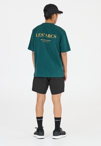 Grön bomullströja med gul text "LES' ARCS" på ryggen, tillsammans med svarta shorts, svarta strumpor och svarta sneakers.