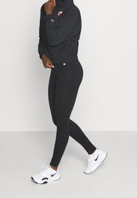 Svart, åtsittande vindjacka med hög krage och logotyp på bröstet, matchad med helsvarta leggings och vita sportskor.