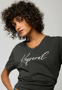 T-shirt gris foncé à col en V fabriqué en tissu doux, affichant un logo en lettres blanches "Kaporal" sur la poitrine. Design décontracté.