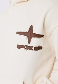 Felpa color crema con un design ricamato marrone irregolare composto da una stella e le lettere "PXD" con bordi sfilacciati.