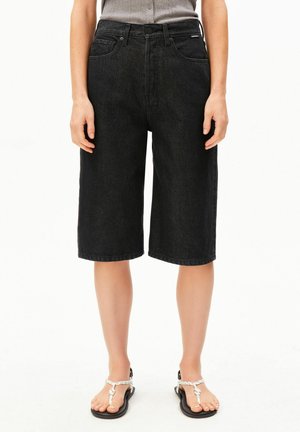 Persoon draagt zwarte denim shorts tot op de knie met voorzakken en een grijze top, staand op voeten met zwarte sandalen met witte bandjes.