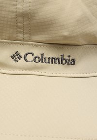 Μπεζ καπέλο με υφασμάτινη υφή, με το λογότυπο της Columbia σε σκούρα ραφή, που παρουσιάζει έναν καθαρό, μοντέρνο σχεδιασμό.