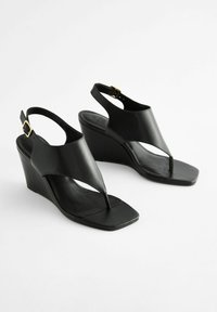 Sorte kilehæle sandaler med en thong-design, fremstillet af glat læder. Funktioner inkluderer justerbar ankelrem med guldspænde og firkantet tå.