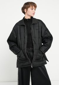 Casaco preto acolchoado com fecho de correr e gola alta. Apresenta uma textura acolchoada e três riscas nas mangas. Combinado com calças pretas.