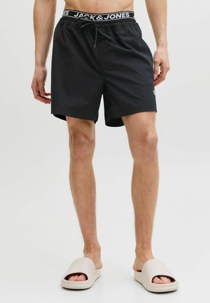 Uomo che indossa pantaloncini da bagno neri Jack & Jones e sandali beige slip-on, in piedi davanti a uno sfondo chiaro e uniforme.