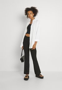 Bílá košile s knoflíky přes černý crop top, spárovaná s černými rozšířenými kalhotami a platformovými sandály. Drží malou černou kabelku.