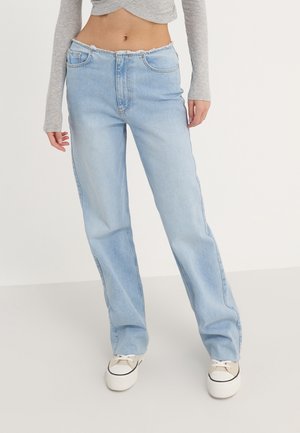 Frau trägt hellblaue, hoch taillierte, gerade geschnittene Jeans, graues langärmliges kurzes Oberteil und weiße Plateausneaker, steht vor einfachem Hintergrund.