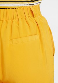 Gula tygbyxor med en samlad elastisk midja och en ficka på sidan; slät textur och enhetlig färg utan mönster.