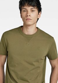 T-shirt in cotone verde oliva con colletto a giro, maniche corte e un logo del marchio discreto sul petto. Texture morbida con vestibilità classica.