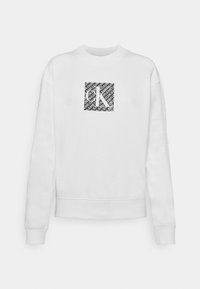 Valkoinen huppari, joka on tehty pehmeästä puuvillasta, ja siinä on ribbaus hihansuissa ja helmassa. Rintakehässä on neliömäinen graafinen kuva, jossa on mustaa toistuvaa tekstiä ja logo.