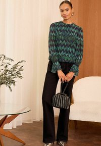 Blusa verde e blu con motivo, maniche plissettate, abbinata a jeans neri a vita alta e una borsa nera strutturata.