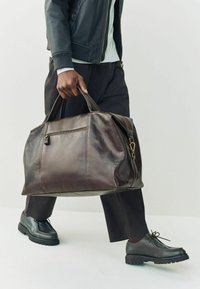 Bolsa de viaje de cuero marrón con cremallera, bolsillo frontal y asas dobles, con una textura suave y detalles de costura sutiles.