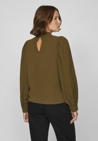 Kvinna med kort brunt hår klädd i en olivgrön blus med lång ärm och detalj i ryggen, tillsammans med svarta byxor, vänd åt sidan.
