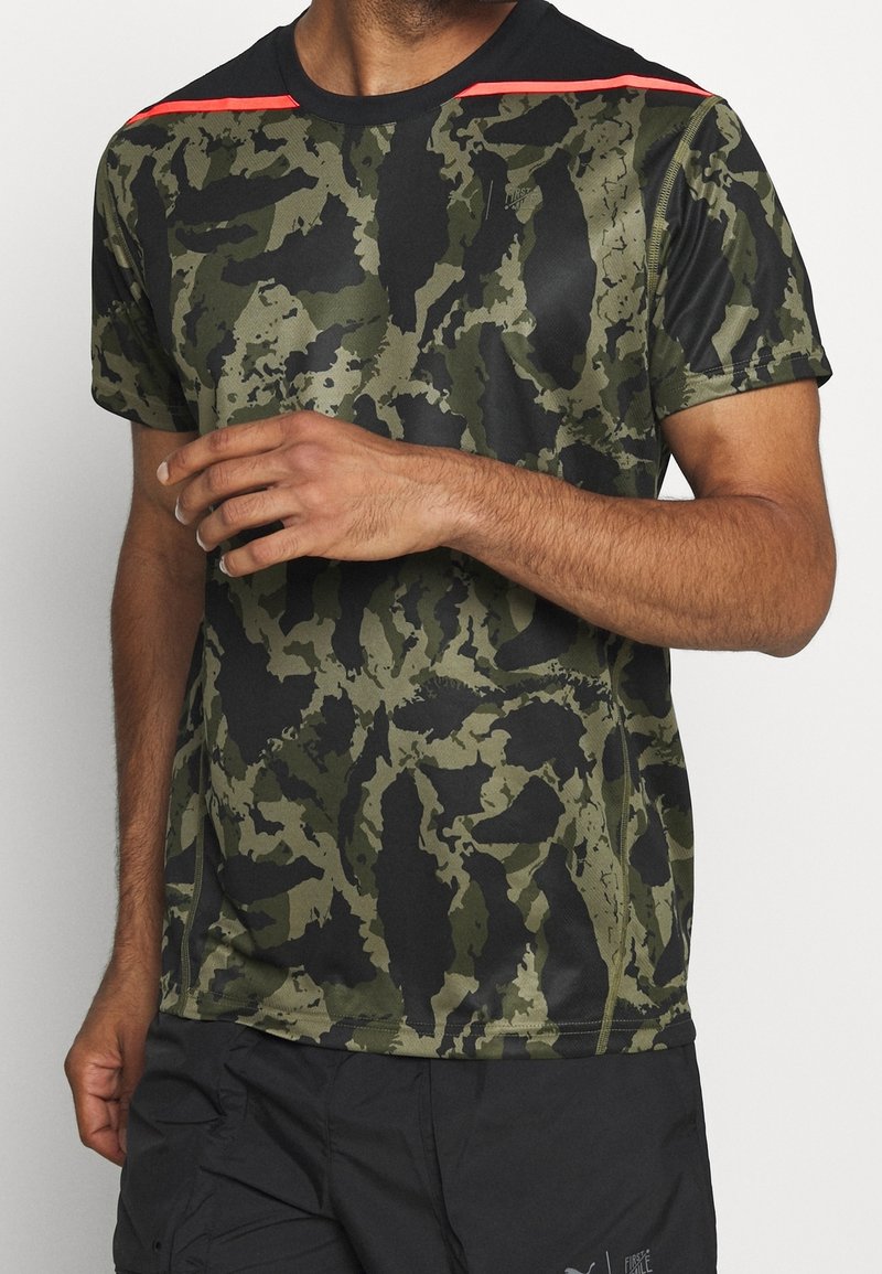 Hombre vestido con camiseta deportiva de camuflaje verde y negro con franjas rojas en los hombros y pantalones cortos negros, con la mano levantada ligeramente.