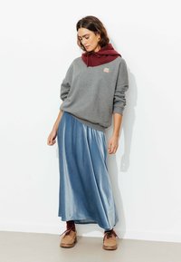 Sweatshirt gris avec une capuche bourgogne, superposé à une jupe maxi en satin bleu ; le mannequin porte des chaussures marron, se tenant contre un mur blanc.