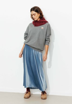 Sweatshirt gris avec une capuche bourgogne, superposé à une jupe maxi en satin bleu ; le mannequin porte des chaussures marron, se tenant contre un mur blanc.