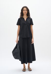 Top corto nero a maniche corte con scollatura a V, abbinato a una gonna nera fluente. Il modello porta una grande borsa di pelle nera e indossa sandali neri.