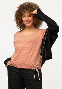Top corto a rayas naranja y crema con mangas cortas, acompañado de una chaqueta negra con cremalleras, dobladillo con cordón y un ajuste relajado. Pantalones negros completan el look.