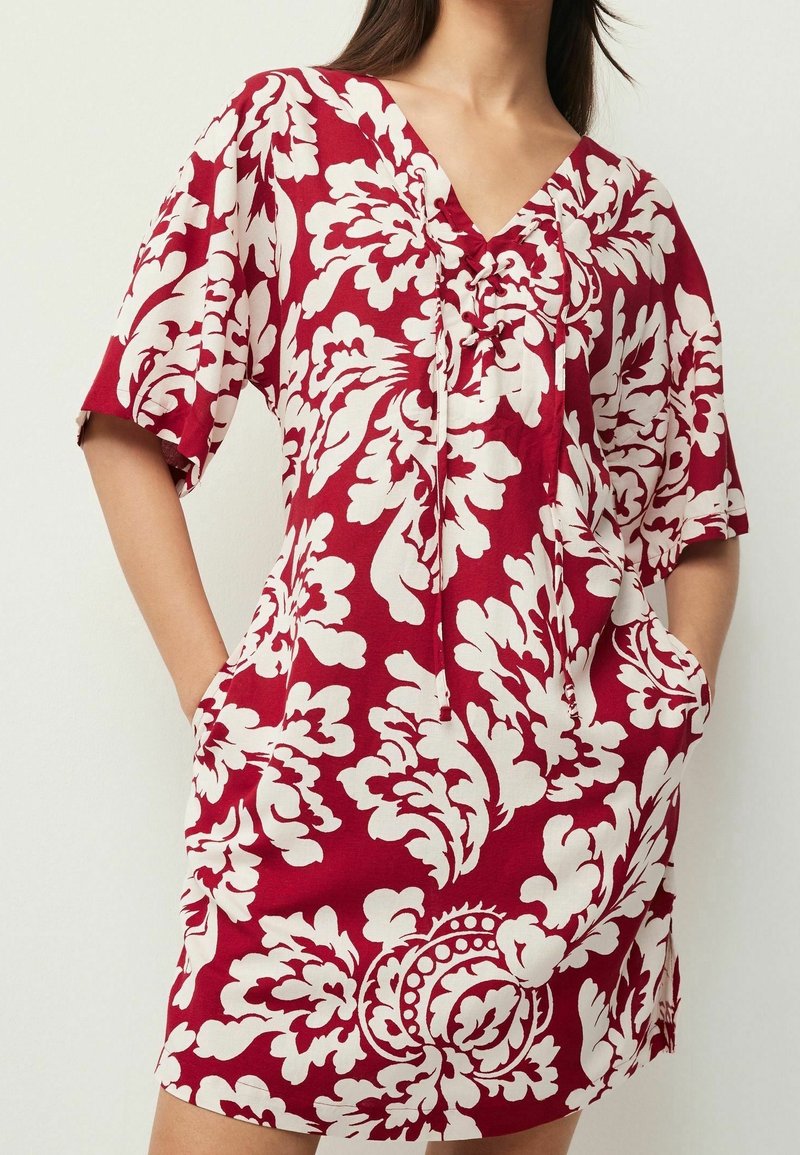 Femme portant une robe rouge à manches courtes avec un motif floral blanc, col en V avec lacets, et les mains dans les poches latérales.