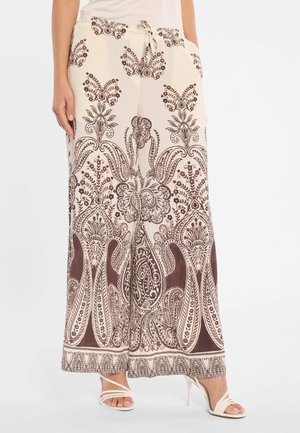 Beige broek met wijde pijpen en ingewikkelde bruine bloemen- en paisleypatronen, een taille met trekkoord, gecombineerd met witte sandalen met hoge hak en bandjes.