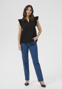 Sort plisseret top med flæseærmer, parret med blå lige ben jeans og sorte højhælede sko. Enkel baggrund, neutral belysning.