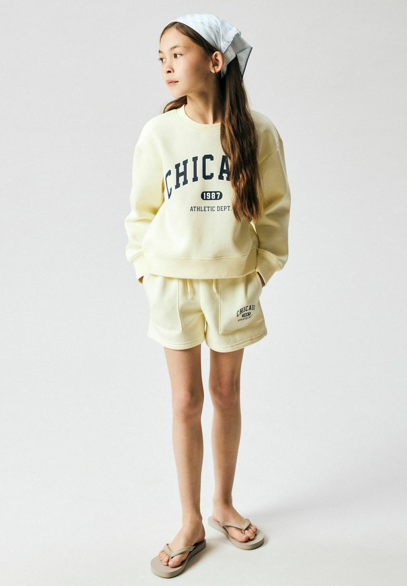 Fille aux longs cheveux portant un sweat-shirt crème et un short assorti avec le texte "Chicago 1987 Athletic Dept.", un foulard blanc sur la tête et des tongs.