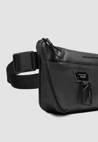 Zwart leren heuptas met een gladde textuur, verstelbare riem, voorvak met ritssluiting en een metalen merklabel als detail.