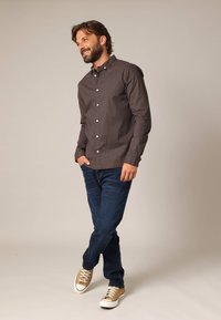 Bruin geruit overhemd met knopen, spijkerbroek en lichtbruine sneakers met witte zolen. Model staat casual met de handen in de zakken.