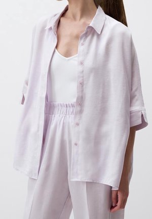 Vrouw draagt een loszittend lichtroze blouse met knopen over een wit tanktopje en bijpassende lichtroze broek met hoge taille tegen een effen achtergrond.