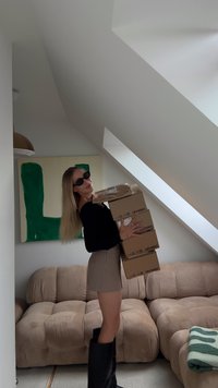 Salmiņi kartona kastēs, uzvelkuši melnus saulesbrilles, melnu ilgu piedurkņu krekls, bēša šorti un melni augsti zābaki, stāv blakus dīvānam.