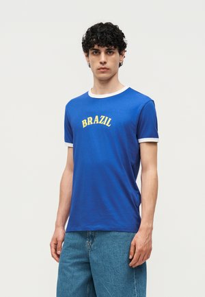 Νεαρός άνδρας με σκουρόχρωμα σγουρά μαλλιά, φορούν μπλε μπλουζάκι με λευκή επένδυση και το «BRAZIL» κίτρινο, συνδυασμένο με φαρδιά μπλε τζιν.