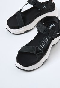 Svarta sportiga sandaler med texturerade svarta remmar, vit mellansula och framträdande varumärkeslogotyp. Öppen tå-design med justerbara stängningar.
