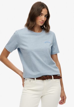T-shirt en coton bleu clair à manches courtes, arborant un logo textuel subtil dans un bleu assorti. Porté avec un jean blanc et une ceinture tressée marron.