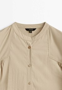 Chemise beige à boutons avec un col rond et des détails cousus le long des coutures d'épaule, posée à plat sur un fond blanc.