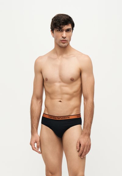 Jeune homme aux cheveux courts et foncés portant un slip noir avec une ceinture orange Emporio Armani sur un fond clair uni.