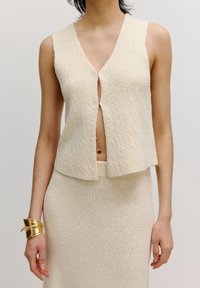 Cremfarbener Strickpullunder mit V-Ausschnitt und offener Vorderseite, der aus einem strukturierten Stoff gefertigt ist. Kombiniert mit einem passenden taillierten Rock. Goldene Armcuff.