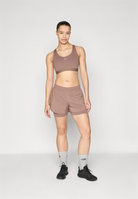 Soutien-gorge de sport marron et shorts assortis en tissu léger. Chaussures de sport noires et chaussettes grises avec logo complètent la tenue.