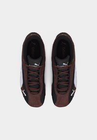 Ζευγάρι αθλητικά παπούτσια Puma με καφέ σουέντ και μαύρο πλέγμα στον άνω μέρος, λευκές ρίγες με λογότυπο και καφέ κορδόνια, σε θέα από πάνω σε λευκό φόντο.