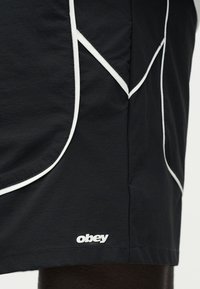 Μαύρο σορτς με λευκές λεπτομέρειες ραφών και μικρό λευκό λογότυπο "obey" κοντά στο στρίφωμα στο αριστερό πόδι.