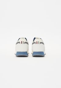 Scarpe da atletica bianche e grigie con suole blu. Presentano una parte superiore in suede e mesh, due sottili strisce rosse sul tallone e una punta arrotondata.