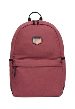 Mochila de tela roja con un diseño redondeado, que cuenta con un bolsillo frontal con cremallera y un parche con el logo. Detalles en negro en la cremallera y las correas.