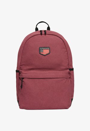 Rode stoffen rugzak met een rond ontwerp, voorzien van een voorvak met rits en een logopatch. Zwarte accenten op de rits en schouderbanden.