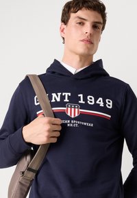 Sötétkék kapucnis pulóver, puha anyagból, nagyméretű "GANT 1949" logóval és pajzs mintával, piros és fehér részletekkel.