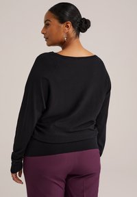 Un pull noir à coupe ample et à encolure large, doté de manches longues et d'un bas côtelé. Le modèle porte un pantalon bordeaux et des boucles d'oreilles en argent.