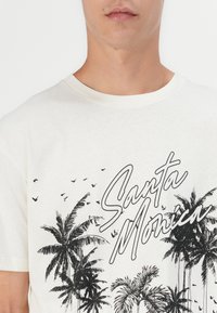 Lys kremet t-skjorte med svarte palmetre-grafikker og teksten "Santa Monica." Klassisk rundhalsdesign med korte ermer.