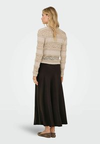 Femme aux longs cheveux blonds portant un haut en dentelle beige à manches longues, une jupe midi noire taille haute et des chaussures plates marron, vue de dos.