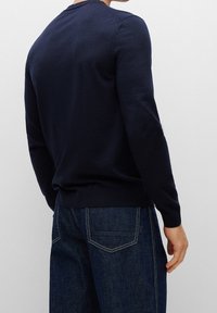 Tmavě modrý pulovr z měkké látky, s dlouhými rukávy, kulatým výstřihem a přiléhavým lemem. Nošený s tmavými džínami.