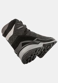 Bota de senderismo negra con una superficie texturizada, paneles de malla y una suela de goma gris con profundas estrías para tracción. Marca Lowa en el costado.