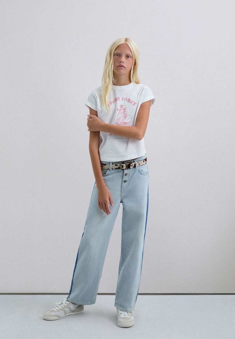 Witte katoenen t-shirt met roze print, gecombineerd met lichtblauwe wijde jeans met blauwe zijkanten, en witte sneakers.